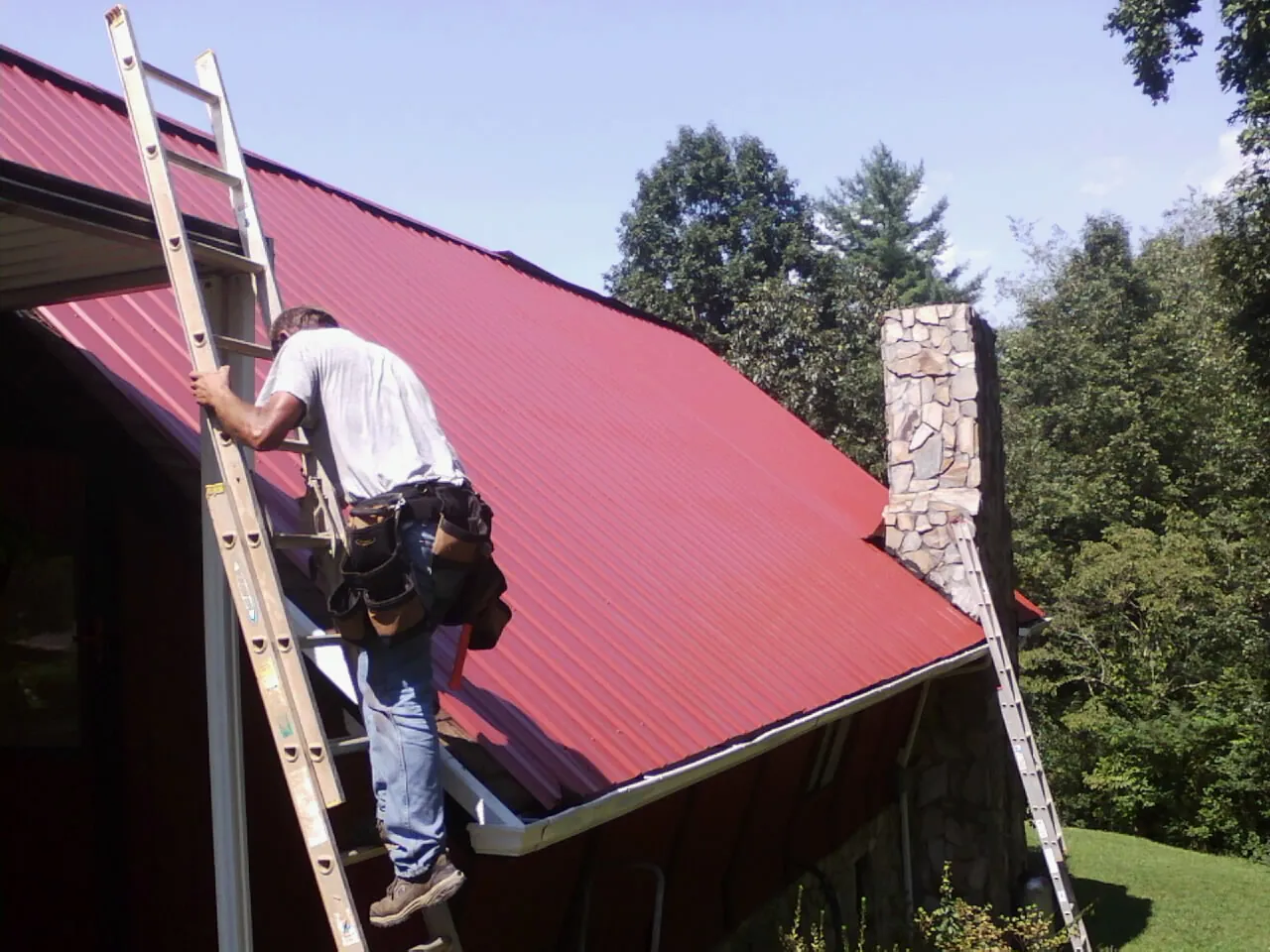 Roofer on a ladder beside a red metal roof and stone chimney — Soffit & Fascia Repair by Roofing Brothers in Hawesville